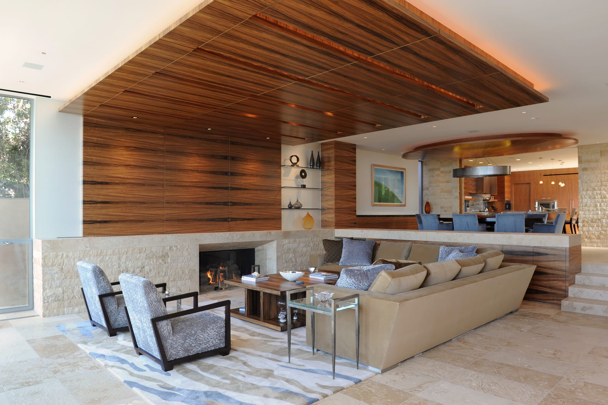 A sunken living room under a floating ceiling of dark wood slats, a fireplace set in stone below