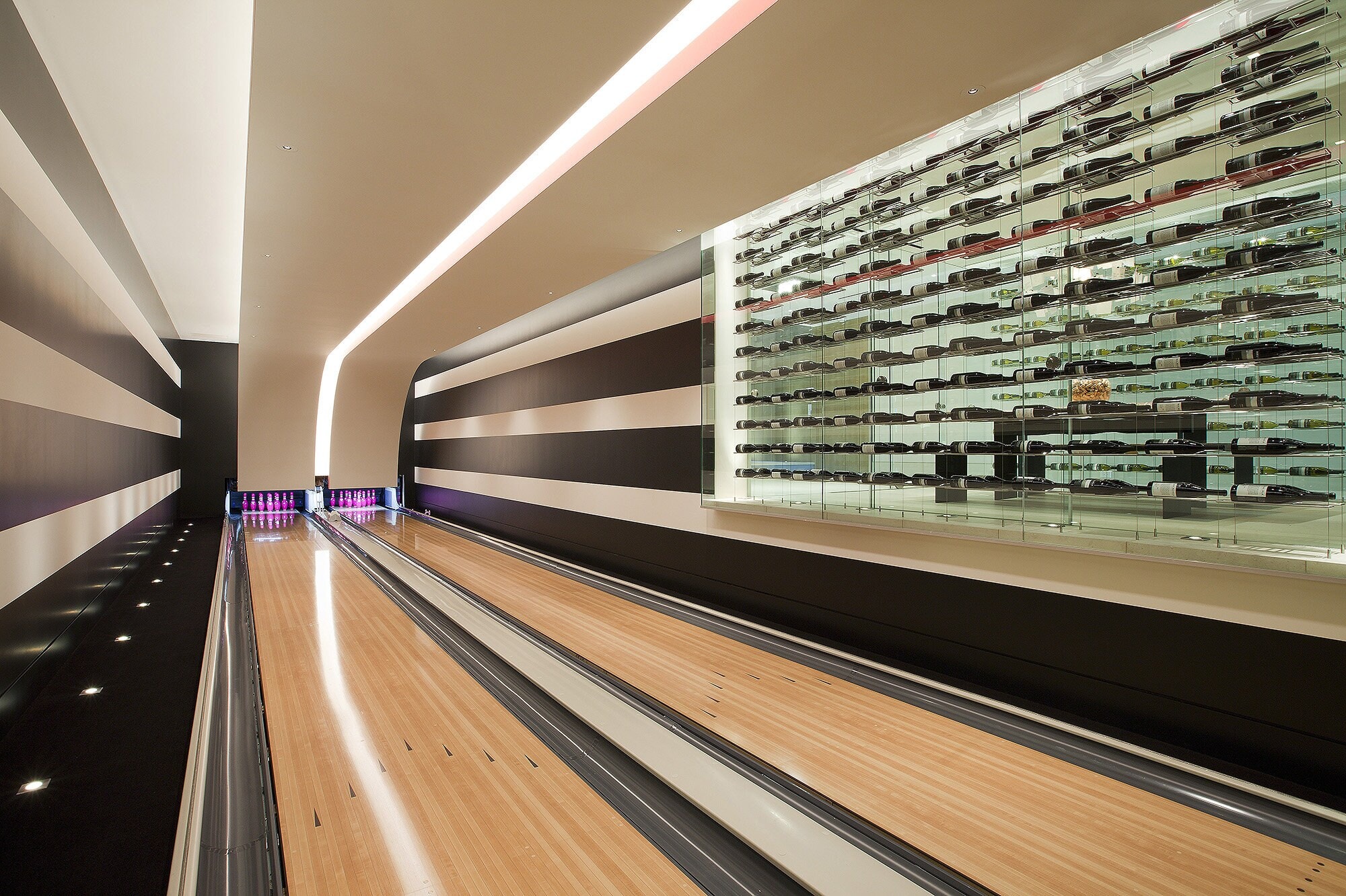 A private bowling lane in pale wood beside a long lit wall of wine