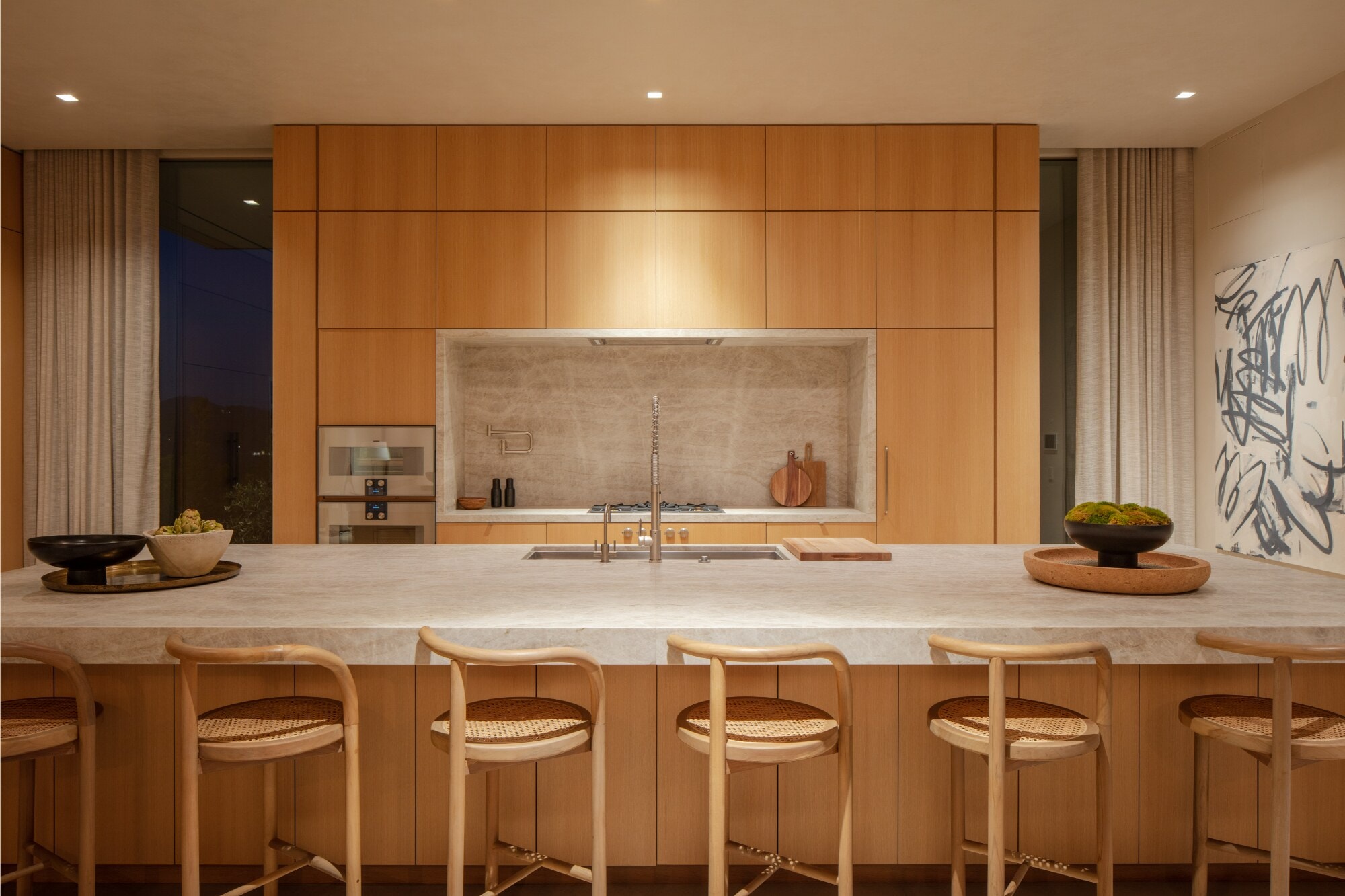A kitchen wall of pale oak cabinetry behind a long stone island