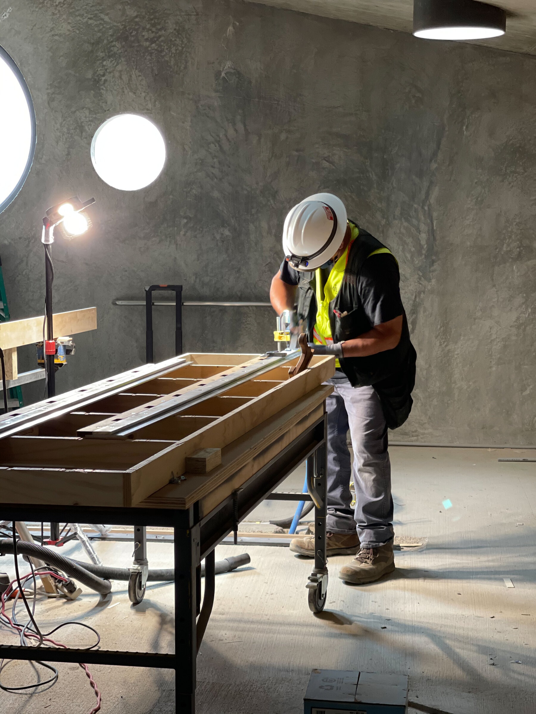 A Miller installer in a hard hat and vest fitting a door on site, lit by a work light beside round windows